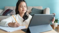 Elementary-aged girl writing in a notebook while working on her tablet during distance learning